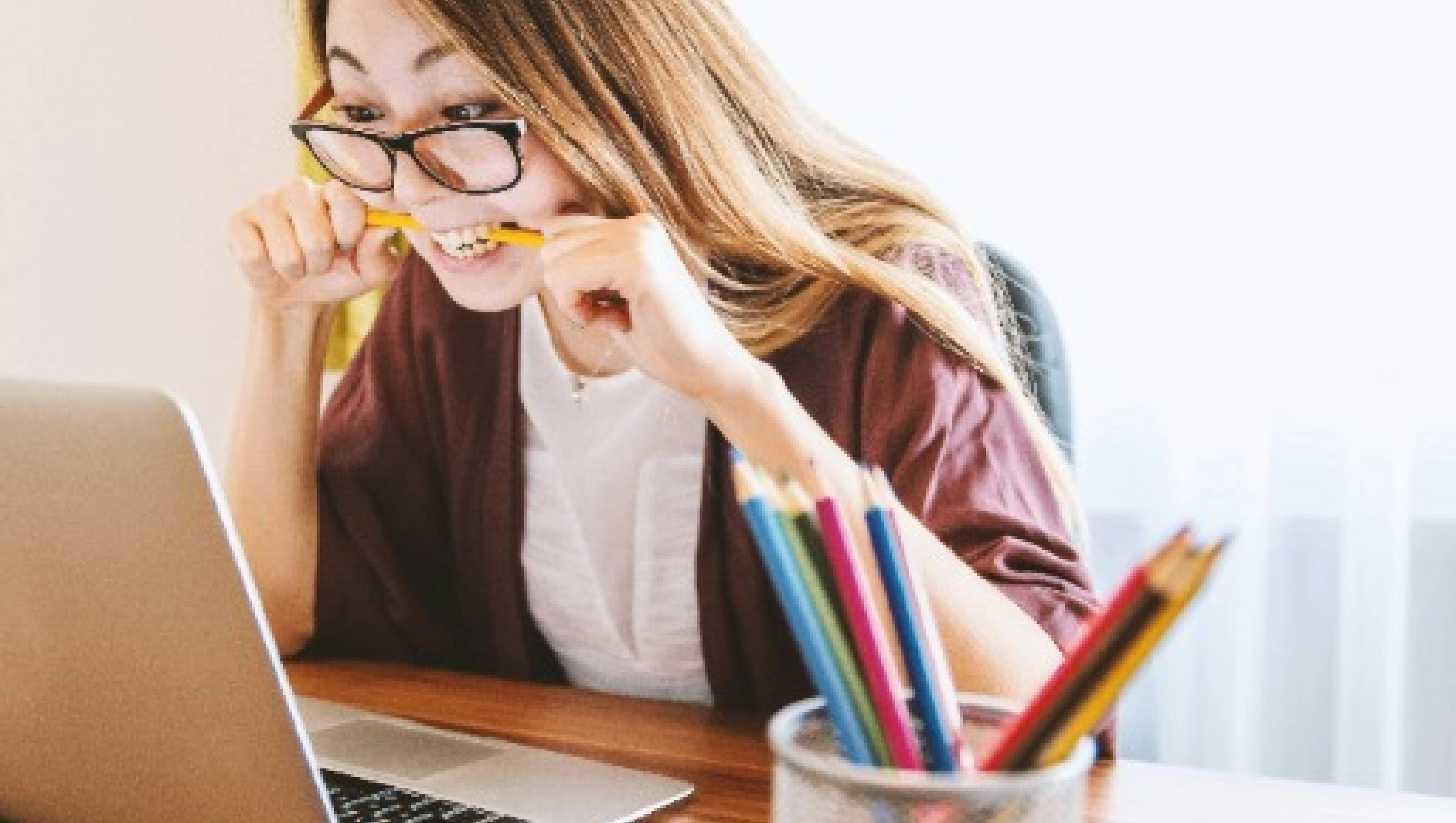 A person with long hair wearing glasses is sitting at a desk, looking at a laptop screen, and biting a yellow pencil in frustration. There is a cup filled with colored pencils in the foreground.