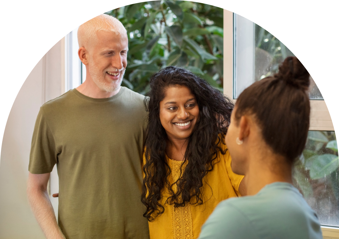 Three adults standing indoors near a window, smiling and talking together. A man with short gray hair stands beside a woman with long dark hair, both looking at another woman in the foreground. Green plants are visible outside the window.