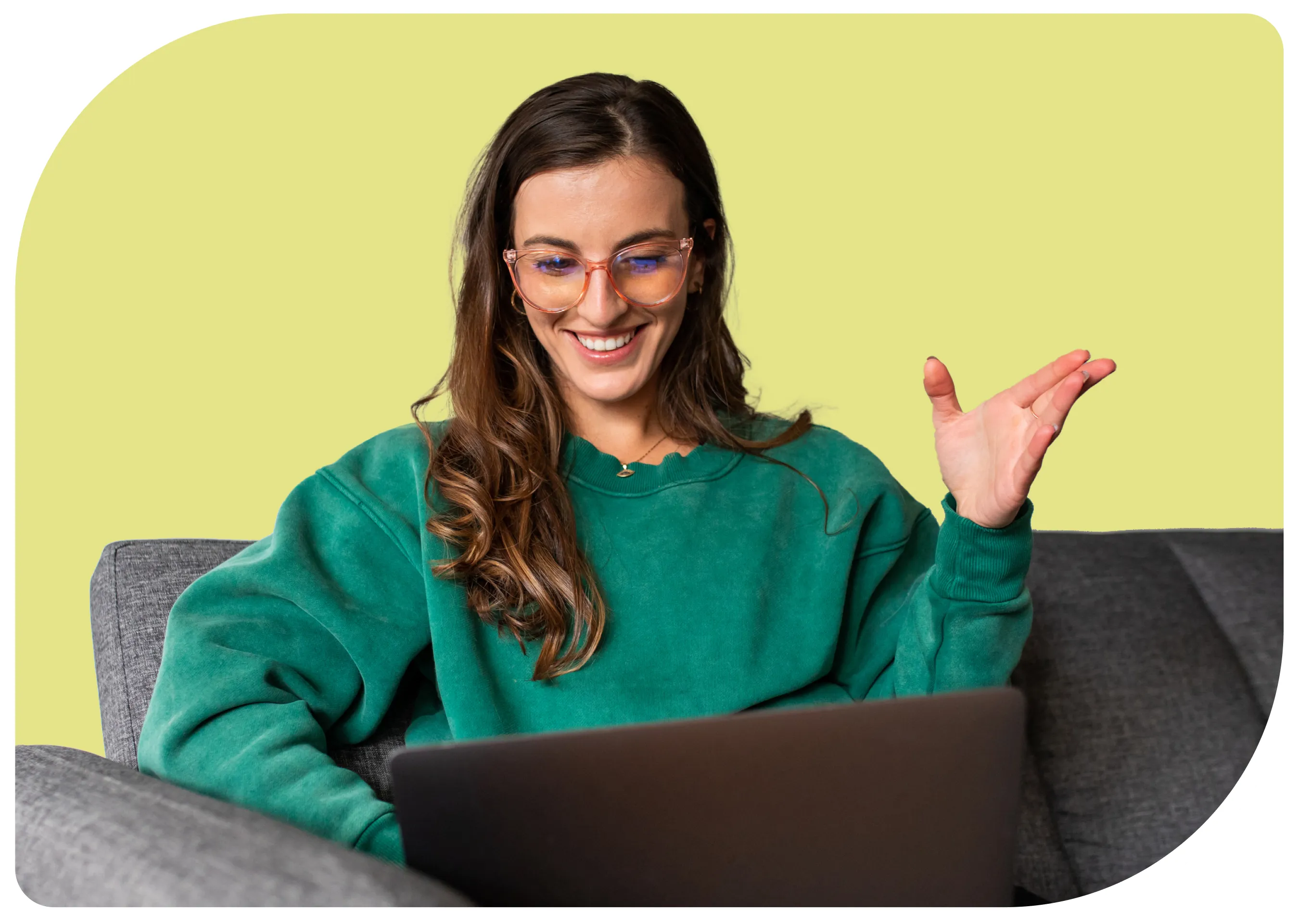 A person with long brown hair and glasses, wearing a green sweater, sits on a gray couch. They are smiling and gesturing with one hand while looking at a laptop in front of them. The background is a solid light yellow color.